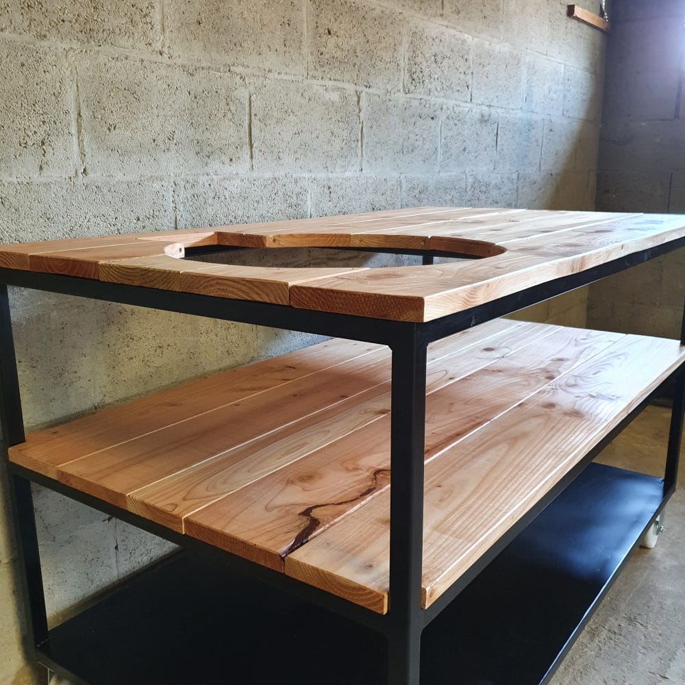 Schuin vooraanzicht van een tafel die bestaat uit 3 verdiepingen. Bovenste 2 zijn van hout en onderste is van staal. Er zit in een gat in het hout vaan een bbq.