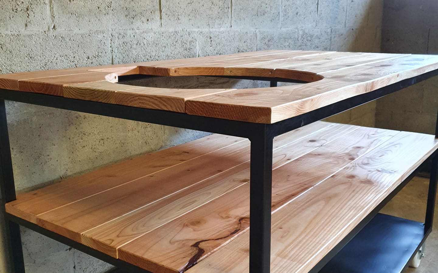 Schuin vooraanzicht van een tafel die bestaat uit 3 verdiepingen. Bovenste 2 zijn van hout en onderste is van staal. Er zit in een gat in het hout vaan een bbq.