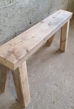 Details van een houten bankje met 4 poten en een dikke houten plank. Staat op de werkplaats voor een betonnen muur, op een betonnen vloer